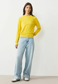 Sárga hosszú ujjú pulóver bordázott kerek nyakkivágással, világoskék, széles szárú farmerral és fehér tornacipővel párosítva, laza szabású és hétköznapi stílusú.