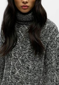 Tykk strikkegenser med høy hals i svart og grått, med intrikate kabelmønstre og teksturert vev, som har en løs og komfortabel passform.