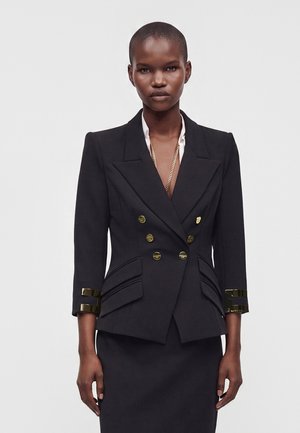Kvinde med kort hår iført en tætsiddende sort dobbeltradet blazer med guldknapper og matchende nederdel mod ensfarvet baggrund.