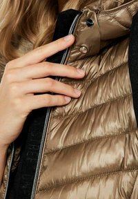 Vadderad jacka med en glänsande, metallisk bronsfoder och svarta detaljer, med dragkedjestängning och tryckknappsdetaljer nära kragen.