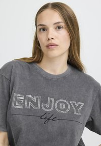 Sivý prebytkový tričko s bielymi kamienkami, ktoré tvoria nápis "ENJOY" a zakrivenú čiaru pod slovom "život". Mäkko, jemne textúrované tkanivo.