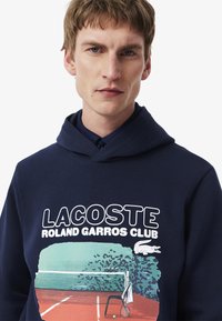Mörkblå hoodie med grafisk tryck av en tennisbana, texten "LACOSTE ROLAND GARROS CLUB" och en krokodil-logotyp. Tillverkas i bomullsmaterial.