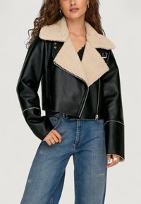 Schwarze Kunstleder-Cropped-Jacke mit einem beigen Lammfellkragen, silbernem Reißverschluss und kontrastierenden weißen Nähten, getragen über blauen Jeans.