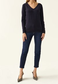 Marineblauer V-Ausschnitt-Pullover aus weichem Material, taillierte Form, kombiniert mit dunklen Jeans. Die Schuhe haben gemusterte Absätze mit spitzen Zehen.