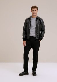 Man som står iklädd en svart läderjacka, grå skjorta, svarta byxor och svarta skor mot en enkel ljus bakgrund.