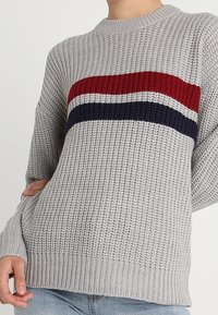 Szary dzianinowy sweter o prążkowanej fakturze, z czerwono-biało-granatowymi poziomymi paskami na wysokości klatki piersiowej. Okrągły dekolt i luźny krój.