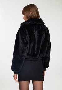Giacca in faux fur nera con colletto alto e design corto. Presenta maniche a sbuffo e orlo a coste, creando una silhouette voluminosa.