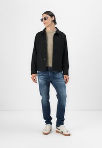 Black button-up jacket, beige knitted sweater, blue jeans, and white sneakers with brown accents. Model wears sunglasses.