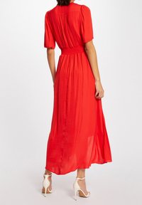 Rood, vloeiende maxi-jurk met korte mouwen en een elastische taille. Heeft een geplooide achterkant en lichte stof. Gaat gepaard met witte hoge hakken.