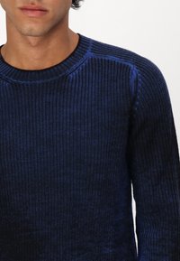 Mörkblå stickad tröja med en strukturerad ribbad mönster. Har en rund halsringning och långa ärmar, och visar upp varierande nyanser av blått.