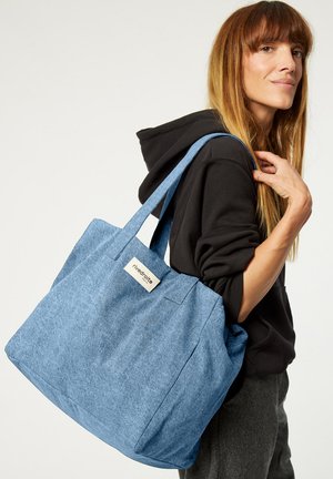 Sac fourre-tout en tissu texturé bleu avec des poignées doubles et une étiquette à l'avant. Grande forme rectangulaire adaptée à divers usages, fabriqué à partir de matériaux durables.