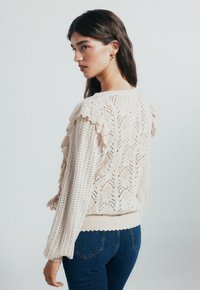 Donna con lunghi capelli scuri che indossa un maglione di pizzo crema con volant e jeans blu, che guarda oltre la spalla su uno sfondo neutro.