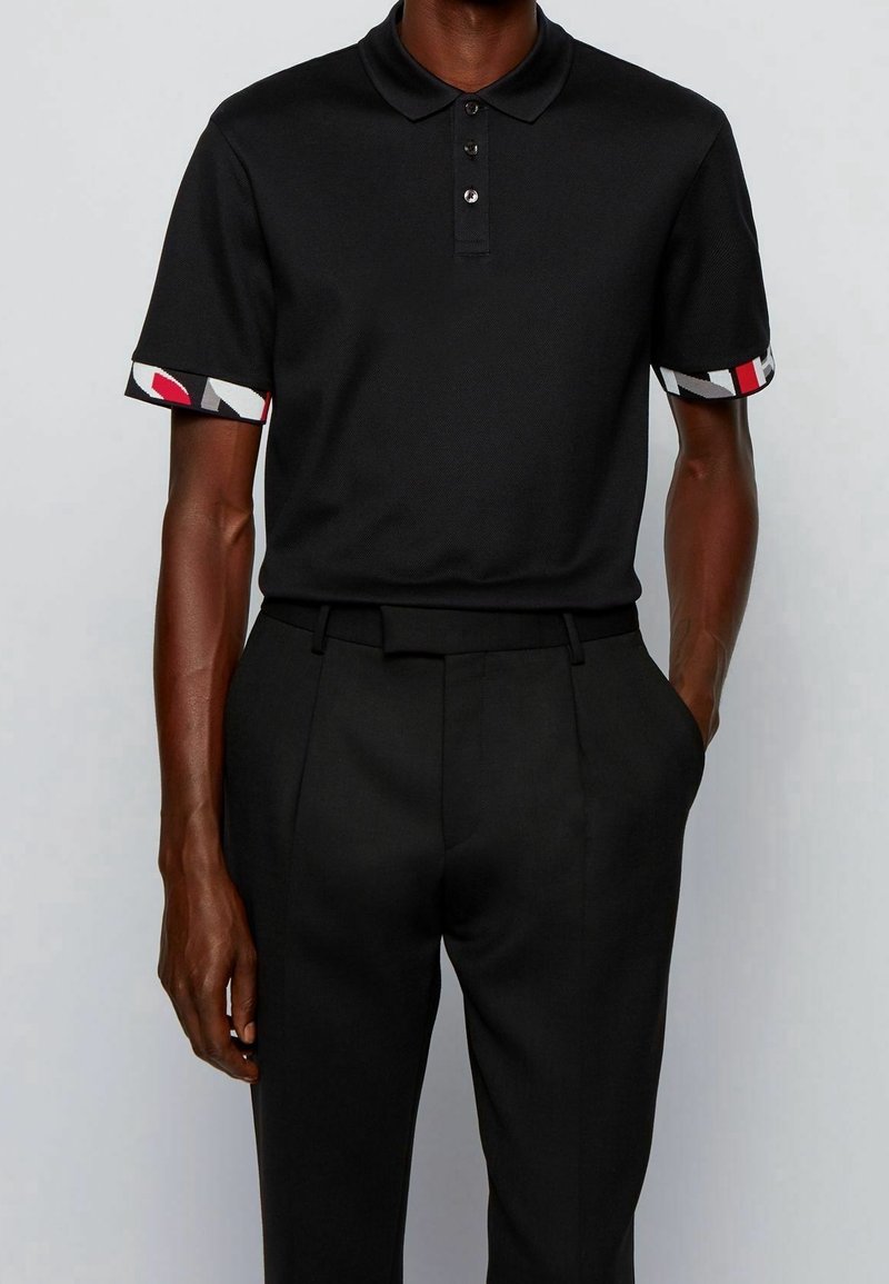Homme portant un polo noir à manches courtes avec des poignets à motifs et un pantalon noir avec ceinture devant un fond uni.
