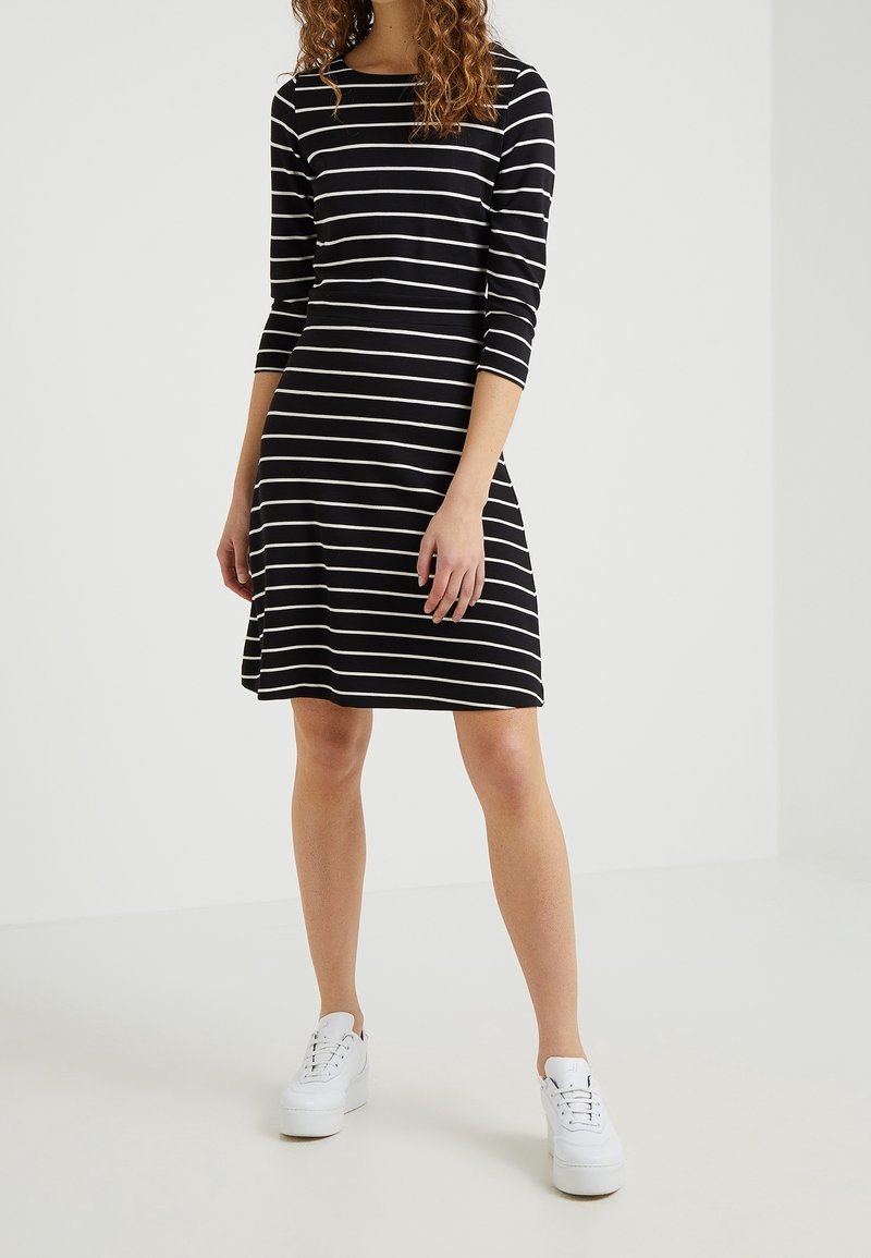 Femme portant une robe rayée horizontale noire et blanche avec des manches trois-quarts et des baskets blanches à plateforme, debout devant un fond uni.