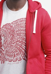 Röd zip-tröja med vita dragsnören, över en ljusgrå t-shirt med en djärv röd grafisk design av ett tribal-motiv.