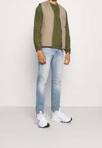 Muž oblečený v světle modrých džínách, bílých teniskách, olivově zelené košili s dlouhým rukávem a béžové prošívané vestě stojí před bílým pozadím.