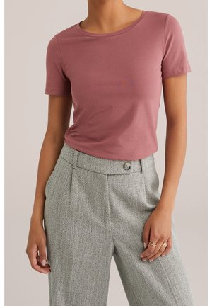 Kobieta ubrana w krótki, różowy, zakurzony t-shirt wpuszczony do wysokich, szarych spodni w jodełkę z guzikami. Pierścionki na prawej ręce.