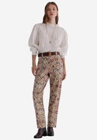Blusa bianca con maniche a sbuffo e dettagli in pizzo abbinata a pantaloni a fantasia floreale in toni terrosi, e una cintura marrone con una fibbia dorata.