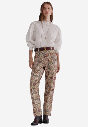 Witte blouse met pofmouwen en kantdetails, gecombineerd met bloemenpatroonbroek in aardetinten, en een bruine riem met een gouden gesp.