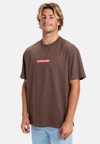 Brun bomulds T-shirt med korte ærmer, der har et rødt "Quiksilver" logo på forsiden. Designet er løst og med en rund halsudskæring.
