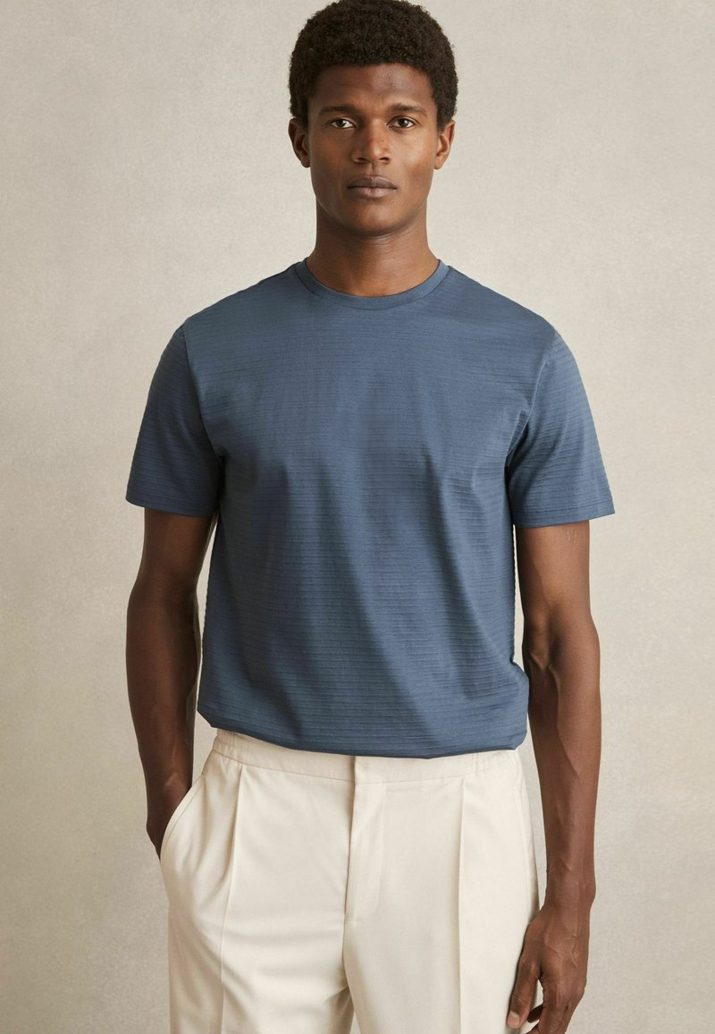 Tricou bleu marin cu mânecă scurtă, cu un model texturat în dungi. Are un guler rotund și o croială lejeră, purtat împreună cu pantaloni de culoare crem.