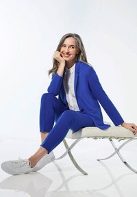 Costume bleu ajusté avec un long blazer et un pantalon fuselé, associé à une chemise blanche à col et des baskets blanches, assis sur une chaise minimaliste.
