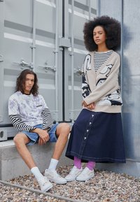 Pull en maille avec manches rayées et motif tie-dye violet, associé à un short en denim. Femme portant une longue jupe en denim foncé et un cardigan à motif.