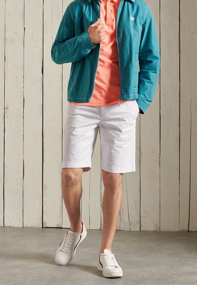 Homme portant une veste turquoise, un polo corail, un short blanc et des baskets blanches, debout contre un mur en bois clair.