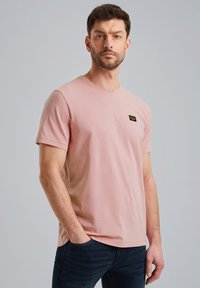 T-shirt s kratkimi rokavi in okroglim izrezom v nežno rožnati barvi iz mehkega bombaža, z majhno črno etiketo na prsih, v kombinaciji s temno modrimi jeans hlačami.