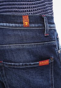 Jean en denim bleu marine avec une étiquette de marque rouge sur la poche arrière et un détail de surpiqûre rouge contrastant le long de la taille.