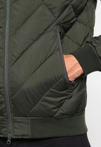 Vadderad jacka i mörkgrön med diamantmönstrad quiltning, framdragkedja, sidofickor och ribbade muddars. Lättvikts syntetmaterial.