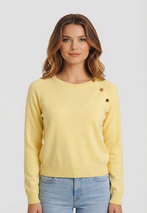 Donna con capelli castani di media lunghezza che indossa un maglione giallo con tre bottoni dorati su una spalla e jeans azzurri.