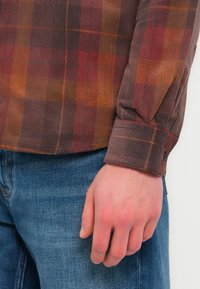 Καφέ και κόκκινο πουκάμισο καρό από υφαντό κοτλέ με ραβδωτή υφή και μανσέτα με κουμπιά, συνδυασμένο με μπλε τζιν παντελόνι.