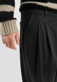 Zwarte geplooide broek met een gladde textuur, gecombineerd met een chunky gebreide trui met zwart en beige strepen. Casual stijl.