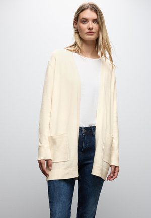 Krämfärgad stickad cardigan med lös passform, ribbade ärmslut och två framfickor. Bärs över en vit topp och matchas med mörkblå jeans.