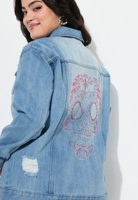 Farkkutakki vaaleansinisellä pesulla, jossa on suuri, monimutkaisesti suunniteltu pääkallo-kuvio pinkeillä timanteilla takana sekä kulutettuja yksityiskohtia.