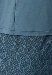 Türkisfarbene Baumwollbluse mit geradem Saum, kombiniert mit einem gemusterten dunkel-türkisen Rock, der Rautenformen und spiralförmige Designs aufweist.
