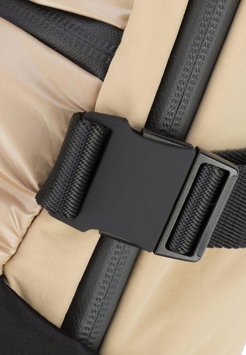 Schwarze Kunststoffschnalle auf beigem Stoff; verfügt über schwarze Riemen und eine strukturierte Oberfläche, mit sichtbaren Nähten und verstellbaren Designelementen.