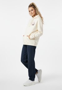 Felpa cremosa con grafica a forma di cuore e tasche, abbinata a jeans testurizzati blu scuro e sneaker bianche. Design semplice, vestibilità casual.