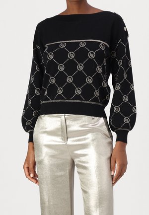 Vrouw draagt een zwarte trui met lange mouwen met lichte geometrische patronen en glanzende zilveren broek tegen een effen achtergrond.