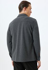 Camisa gris oscuro de tela texturizada, con cuello, mangas largas y puños abotonados. Diseño sencillo y funcional.