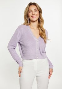 Cardigan lavorato a maglia lavanda con scollo a V, maniche lunghe e chiusura con bottoni. Presenta una sottile texture e un orlo corto, abbinato a pantaloni bianchi.
