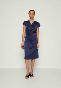 Mornarsko modra satenska obleka z zavihkom, kratkimi rokavi in dolžino do kolen, ter subtilnim leskom. Črna torbica in sandali z odprtimi prsti dopolnjujejo videz.