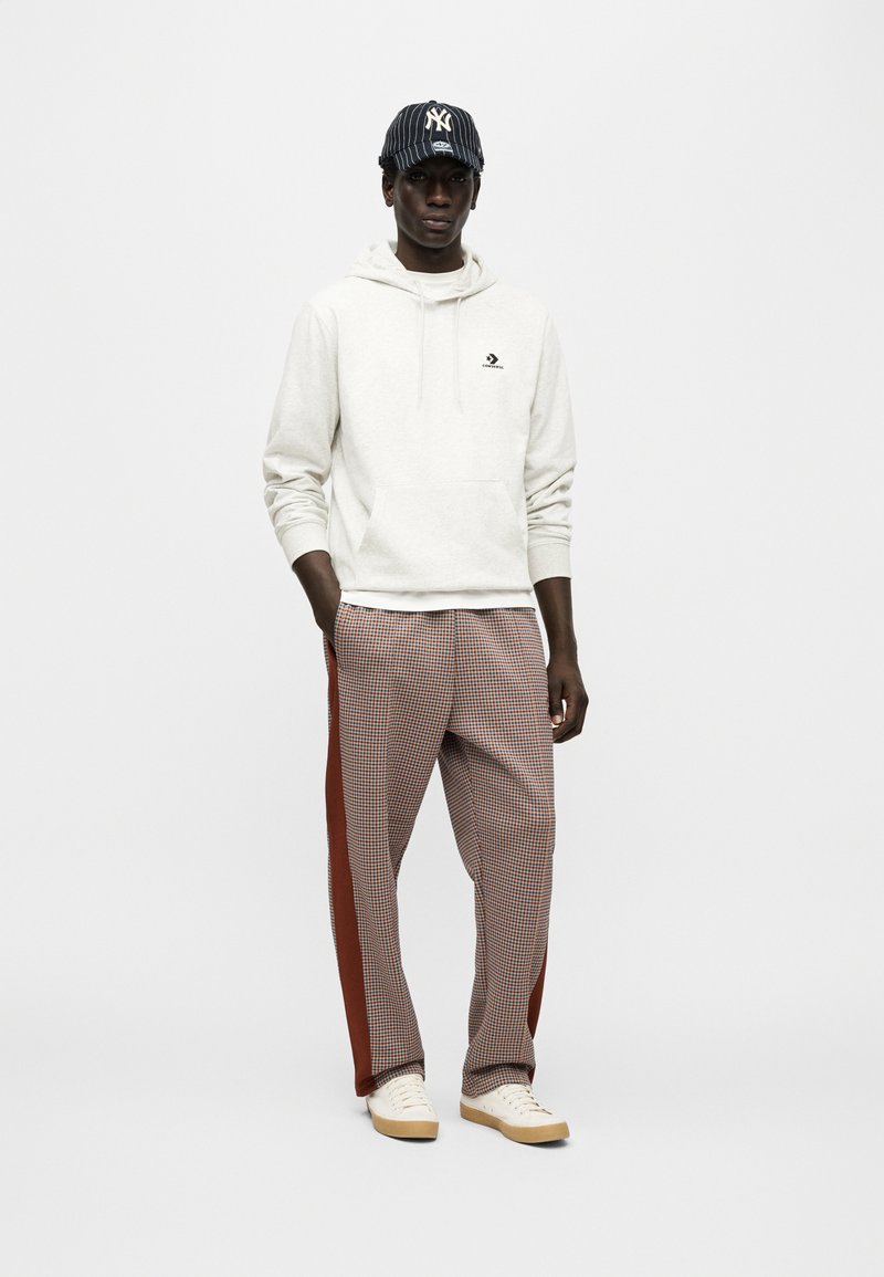 Sweat à capuche crème avec poche avant, pantalon à carreaux avec rayures latérales rouges, et casquette marine à fines rayures blanches. Baskets blanches complètent le look.