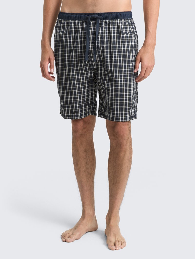 Mand iført navy blå og hvid ternede shorts med snor i taljen, står barfodet imod en enkelt lys baggrund.