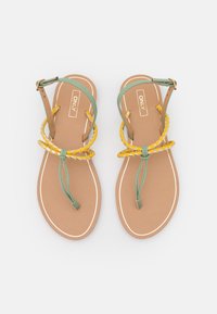 Un paio di sandali piatti con suole color tan, cinghie per le dita verdi, cinghie per le caviglie intrecciate gialle e bianche, e fibbie regolabili su uno sfondo bianco.