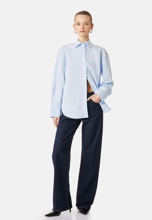 Vrouw draagt een lichtblauw oversized overhemd met knopen, donkere wijde jeans, zwarte puntige hakken en creolen, poseert met één hand in de zak.