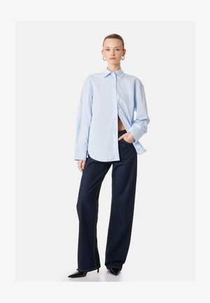 Femme portant une chemise oversize bleu clair boutonnée, un jean large foncé, des talons noirs pointus et des boucles d'oreilles anneaux, posant avec une main dans la poche.