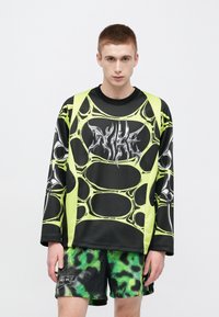 Μαύρο πουκάμισο με μακριά μανίκια και έντονο σχέδιο από φωσφορίζον πράσινο πλεκτό ιστό αράχνης, με στυλιζαρισμένο λογότυπο στο στήθος. Συνδυάζεται με σορτς σε πράσινο και μαύρο μοτίβο.