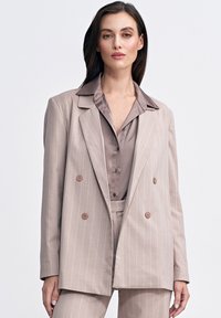 Beżowa marynarka w paski z podwójnym zapięciem, ostrymi klapami i dużymi brązowymi guzikami, nałożona na satynową bluzkę w jasnobrązowym kolorze.